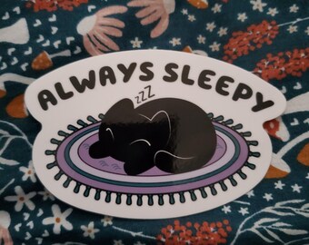 May include: An oval sticker with the words "ALWAYS SLEEPY" in black. It depicts a sleeping black cat on a purple and white rug. The sticker has a scalloped edge.