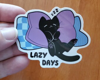 May include: A die-cut sticker featuring a black cat sleeping on a purple pillow with blue checkered accents. The cat has closed eyes and a small smile, with the text 'zzz' above its head and the words 'LAZY DAYS' printed in black below the cat.