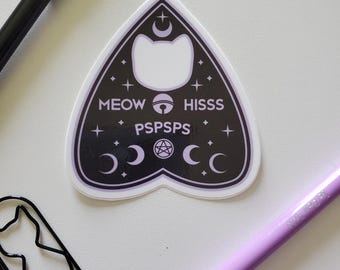 May include: A black and white cat-themed Ouija board sticker with the words "MEOW", "HISSS", and "PSPSPS" printed on it. The sticker has a cat-shaped cutout and crescent moon and star designs. A black pen, a cat-shaped paperclip, and purple pens are also visible.
