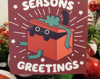 May include: A festive greeting card with a maroon backdrop, displaying "SEASONS GREETINGS" in white. It features a cartoon black cat emerging from a red gift box, adorned with a green bow. White starbursts and stars embellish the design.
