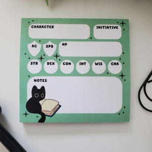 May include: A square, light green notepad with black text and a black cat illustration. The notepad has sections for character stats, initiative, and notes. The cat is reading a book. The notepad is decorated with small star accents.