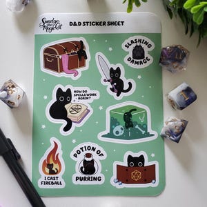 May include: A green D&D sticker sheet with black cat illustrations and phrases such as "Slashing Damage" and "Potion of Purring." The sheet includes a treasure chest, a sword, and a fireball design. Dice are scattered around the sheet.