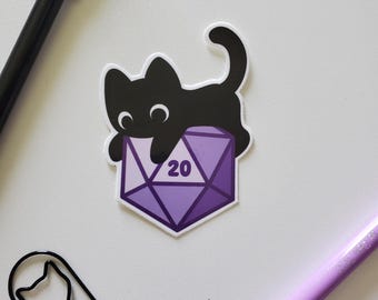 May include: A black cat sticker on a purple and white D20 die, showing the number 20. The sticker is on a white surface, with a black pen, a cat-shaped paperclip, and a purple pen nearby.
