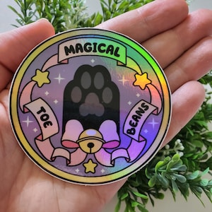 May include: A holographic sticker with a cat paw print design. The sticker features the words "Magical Toe Beans" on banners, with stars and a bow. The paw print is black, and the overall design has a pastel color scheme.