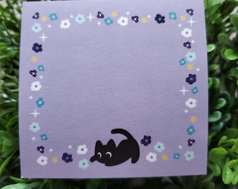 May include: A square, lilac sticky note with a border of small flowers in white, blue, and yellow. A black cat illustration is in the lower right corner. The note is surrounded by green foliage.