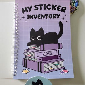 May include: A spiral-bound notebook with the text "MY STICKER INVENTORY" and a black cat illustration. The cover is light purple with a grid pattern. Two round stickers are also visible, one with a cat holding a knife and the other with a rainbow design.