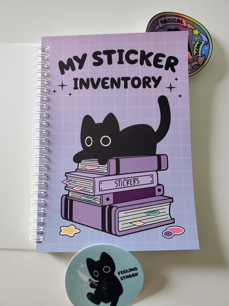 May include: A spiral-bound notebook with the text "MY STICKER INVENTORY" and a black cat illustration. The cover is light purple with a grid pattern. Two round stickers are also visible, one with a cat holding a knife and the other with a rainbow design.