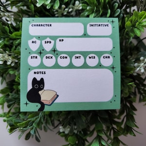 Op de afbeelding: Een mintgroen notitieblok voor een personageblad met witte vakken voor statistieken en notities. Het notitieblok heeft een illustratie van een zwarte kat die een boek leest. De woorden "CHARACTER", "INITIATIVE" en andere RPG-termen staan erop.