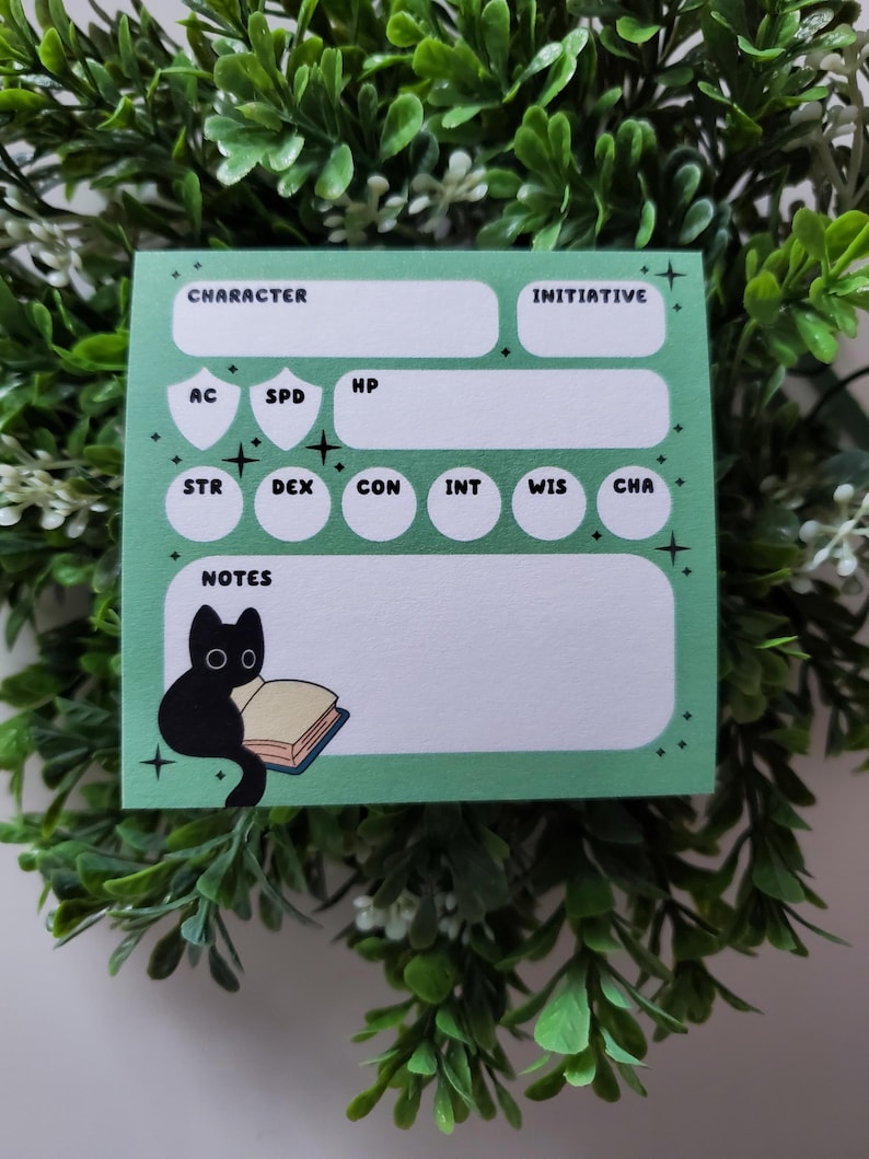 Op de afbeelding: Een mintgroen notitieblok voor een personageblad met witte vakken voor statistieken en notities. Het notitieblok heeft een illustratie van een zwarte kat die een boek leest. De woorden "CHARACTER", "INITIATIVE" en andere RPG-termen staan erop.