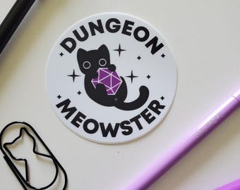 May include: A white circular sticker featuring the words "DUNGEON MEOWSTER" around a black cat holding a purple die. A black cat-shaped paperclip, a black pen, and two purple pens are also visible.
