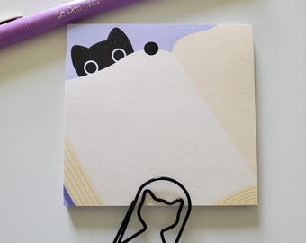 May include: A square sticky note featuring a black cat illustration, a purple pen, and a black cat-shaped paper clip. The pen is labelled "Le Pen". The sticky note has a light purple and beige design.