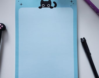 May include: A pale blue notepad featuring a black cat illustration at the top. The notepad has a light blue interior and a darker blue border. A black pen and a purple pen are visible.