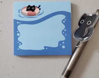 May include: A square sticky note pad featuring a light blue centre with dark blue wavy borders and a black cat floating in a pink and white inner tube at the top, accompanied by a silver pen with a black cat-shaped clip attached to the barrel.