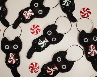 May include: Black felt cat decorations with white eyes and a candy accent. The decorations have a loop for hanging and are surrounded by red and green peppermint sweets. The cat decorations are a festive decoration.