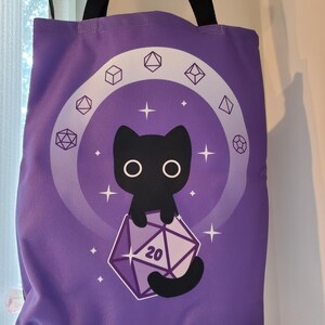May include: A purple tote bag with black handles featuring a graphic of a black cat holding a twenty-sided die with the number 20 printed on it. The design includes a circular arrangement of various polyhedral dice shapes and white stars.
