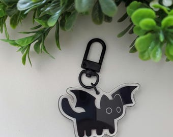 May include: A black cat with bat wings keyring. The keyring has a black clasp and a clear outline. The cat has two round eyes and a tail. The background is white with green foliage.