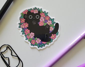 May include: A sticker featuring a black cat surrounded by pink and purple flowers. The cat is lying on its back. A black pen, two purple pens, and a cat-shaped paperclip are also in the image.