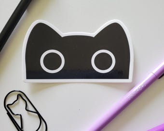 May include: A black cat-shaped sticker with white eyes and a white border is surrounded by stationery. A black pen with a cat-ear top, a black cat-shaped paperclip, and two purple pens are arranged on a white surface.