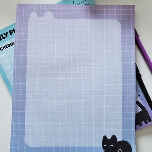 May include: A cat-themed notepad with a blue and purple gradient background and a grid pattern. The notepad features a black cat silhouette and a cat-shaped paperclip. A purple pen is also visible.