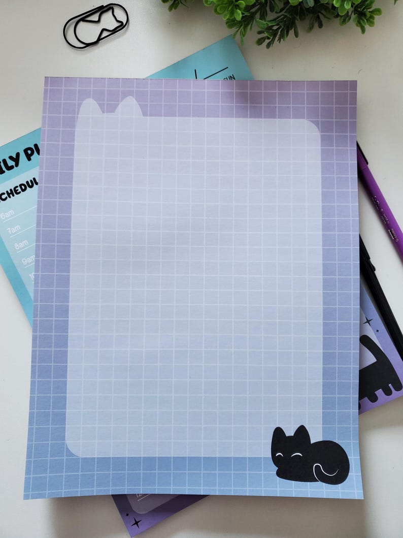 May include: A cat-themed notepad with a blue and purple gradient background and a grid pattern. The notepad features a black cat silhouette and a cat-shaped paperclip. A purple pen is also visible.