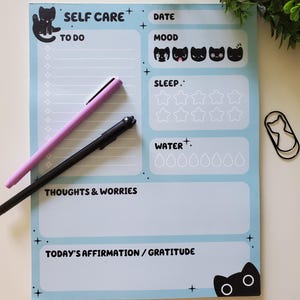 May include: A light blue self-care planner with black cat illustrations and text. Features sections for to-do lists, mood tracking, sleep, water intake, thoughts, and affirmations. Includes a pink and black pen and a cat-shaped paperclip.