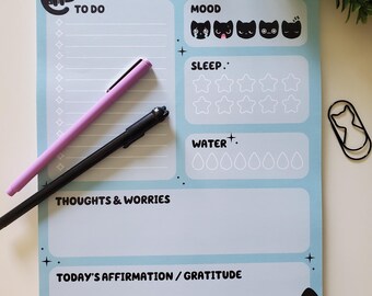 May include: A pale blue self-care planner with black cat illustrations and text. Includes sections for to-do lists, mood tracking, sleep, water intake, thoughts, and affirmations. Features a pink and black pen and a cat-shaped paperclip.