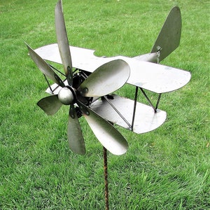 May include: A metal garden sculpture of a biplane with a propeller. The sculpture is painted silver and has a long, thin metal pole for mounting.
