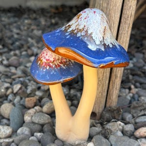 May include: Two ceramic mushroom figurines with blue and white speckled caps and beige stems. The mushrooms are sitting on a bed of small gray and brown stones.
