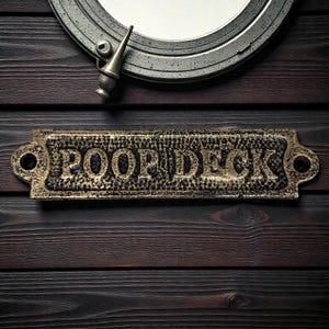 May include: A decorative brass sign with the words "POOP DECK" in raised lettering, mounted on a dark wood surface. Above the sign is a metal porthole with a silver finish and a small, pointed metal fixture.