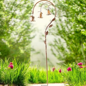 May include: A bronze-colored outdoor garden stake with three bell-shaped lights. The stake has a curved top with leaf and vine details. The lights are amber-colored and hang from the curved top. The stake is placed in a garden setting with green grass and flowers.
