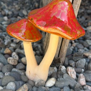 May include: Two ceramic mushrooms with red and gold glaze. The mushrooms are sitting on a bed of small gray and brown rocks.