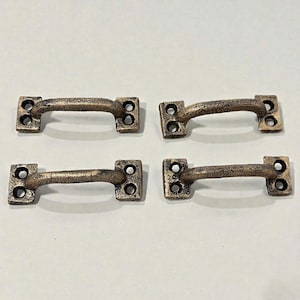 May include: Four antique bronze-colored cabinet pulls. Each pull has a curved handle and two rectangular mounting plates with two holes each. The hardware has a distressed finish, suggesting an aged appearance.