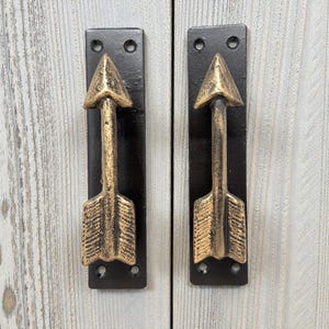 May include: Two cabinet pulls with an arrow design. The arrow is gold-colored with a textured finish, mounted on a black rectangular plate. The pulls are attached to a light-colored, wood-grain surface.