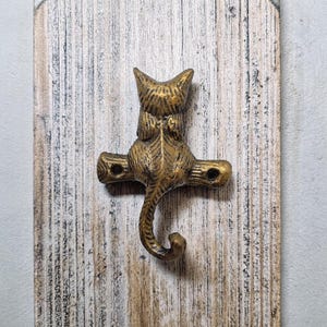 May include: A decorative brass cat hook mounted on a weathered wooden board. The cat hook features a detailed design with a tail that curves downward to form the hook. The wood has a distressed, rustic appearance.