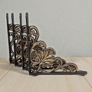 May include: Set of five black and gold decorative shelf brackets. Each bracket features an ornate, floral design with a dark black base and gold accents. The brackets are made of cast iron and are designed to be mounted on a wall.