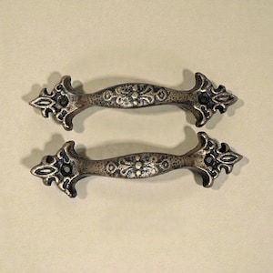 May include: Two antique bronze-colored drawer pulls with ornate floral and arrow-shaped designs. The hardware is made of metal and features a distressed finish, suitable for cabinets or furniture. Each pull measures approximately 6 inches in length.