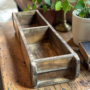 May include: A rectangular, two-compartment wooden brick mold with a weathered, dark brown finish. The structure features visible metal fasteners along the top edges, creating a distinct, segmented open container with a rustic, aged appearance.