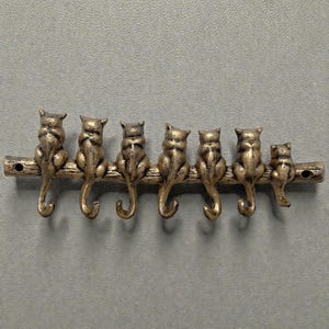 May include: A decorative wall hook featuring seven cat figures sitting on a branch. The hooks are gold-colored and the cats are facing away from the viewer. The piece is mounted on a gray background.