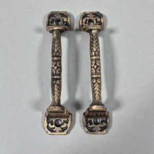 May include: Two antique bronze-colored cabinet pulls. Each pull features a rectangular base with a decorative floral design, a detailed stem, and a top with a circular hole. The pulls are symmetrical and have a vintage aesthetic.
