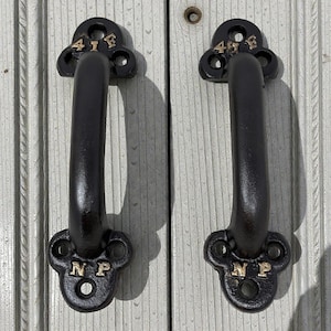 May include: Two black cast iron door handles with a curved design. The handles have decorative ends with the letters "41F" and "NP" in gold. The handles are mounted on a weathered, light-colored wooden surface.