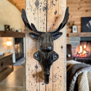 May include: A black cast iron moose head wall hook mounted on a weathered wooden post. The moose head features detailed antlers and a single hook below. The rustic design adds a touch of woodland charm to any space.