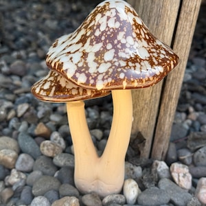 May include: Two ceramic mushrooms with brown and white speckled caps and a light brown stem. The mushrooms are sitting on a bed of small gray and brown rocks.