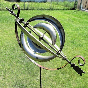 May include: A metal garden sculpture with a circular design. The sculpture is made of silver and brown metal and has a pointed arrow at the top. The sculpture is mounted on a metal pole.