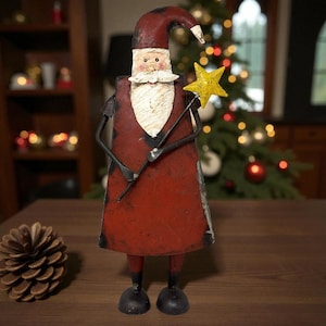 May include: A rustic metal Santa Claus figurine with a red coat, black boots, and a long white beard. The Santa holds a yellow star wand. The figurine is on a wooden surface, with a pine cone in the foreground and a Christmas tree in the background.