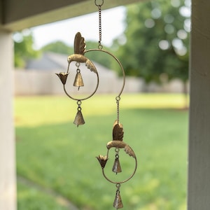 May include: A decorative wind chime featuring two hummingbirds crafted from bronze-colored metal. Each hummingbird is perched on a circular frame with a flower and a small bell. The chimes hang from a chain, creating a gentle, melodic sound in the breeze.