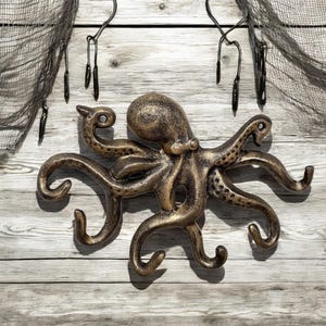 May include: A gold-colored metal octopus wall hook with eight tentacles that are shaped into hooks. The octopus is mounted on a white wooden wall.