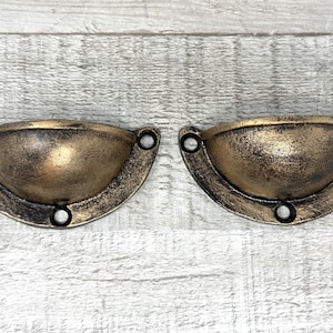 May include: Two antique-style drawer pulls with a bronze finish. Each pull is a half-circle shape with a rounded bowl and three holes for mounting. The pulls have a distressed, aged appearance.