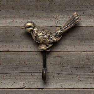 May include: A decorative wall hook featuring a bird design. The bird is cast in a gold and black finish, perched atop a dark metal hook. The hook is mounted on a wooden surface.