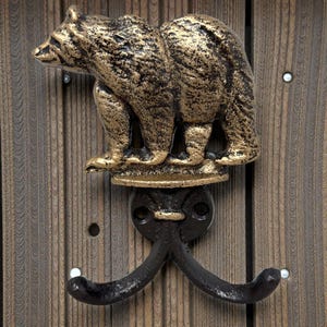 May include: A decorative wall hook featuring a bear design. The bear is painted in gold and black, mounted on a black double hook. The hook is attached to a wooden surface.