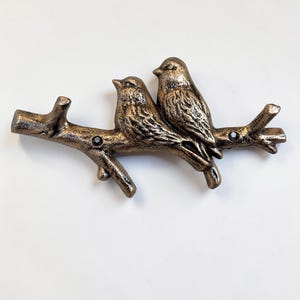 May include: Bronze-colored metal sculpture of two birds perched on a branch. The birds have detailed feather textures and are positioned side-by-side. The branch has a textured, rustic appearance, with small black accents. The sculpture is likely a decorative item.